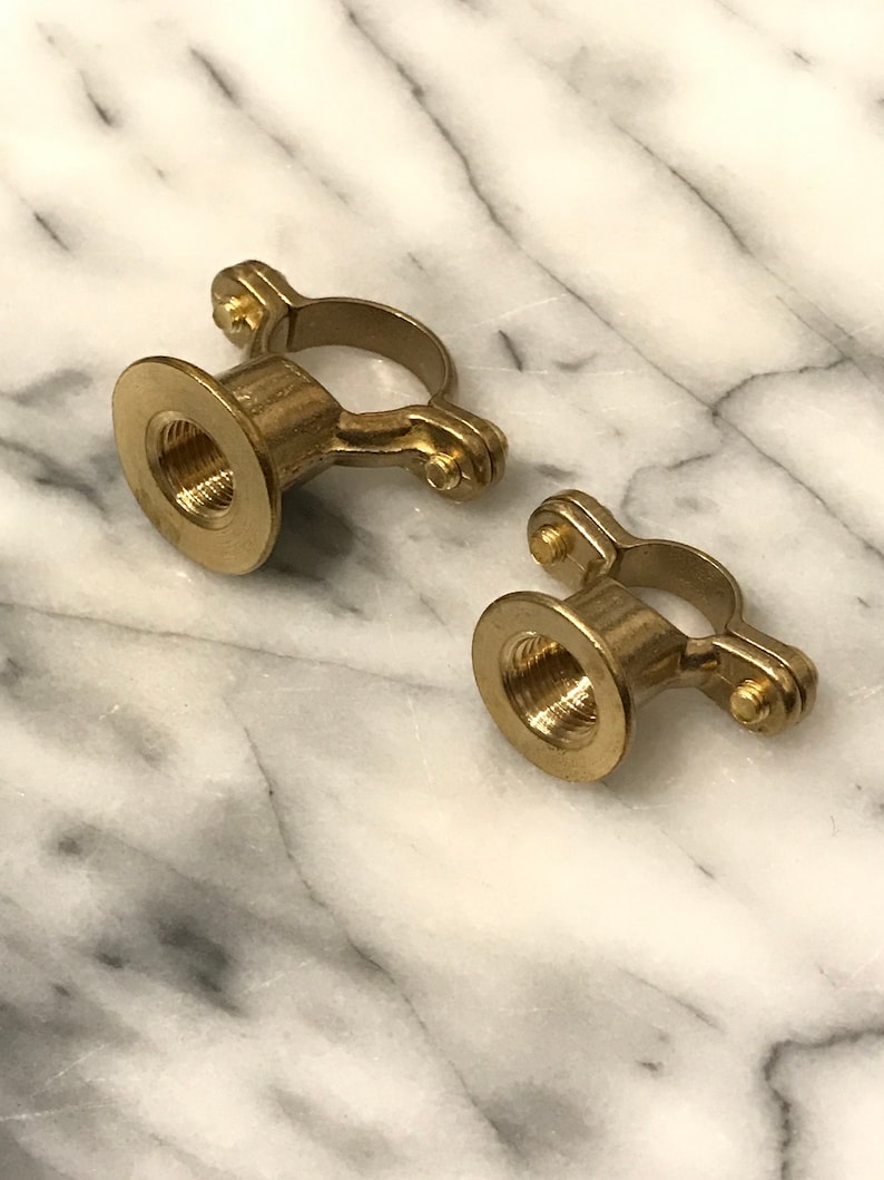 May include: Two brass pipe clamps with a threaded interior and a circular ring. The clamps are made of a shiny, golden metal and are set against a white and grey marble background.