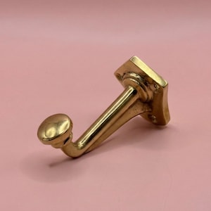 Könnte beinhalten: Ein messingfarbener Wandhaken mit quadratischer Basis und einem gebogenen Arm, der in einem abgerundeten Knauf endet. Der Haken ist vor einem zartrosa Hintergrund platziert, der sein Vintage-Design hervorhebt. Dieser dekorative Haken eignet sich zum Aufhängen von Gegenständen.