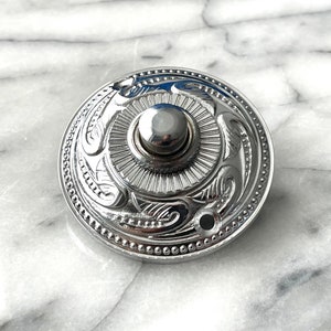 Op de afbeelding: Een zilveren deurbel met een decoratief ontwerp. De deurbel heeft een ronde, vlakke voorkant met een verhoogde knop in het midden. Het ontwerp heeft een zonnestralenpatroon en zwierige bloemmotieven.