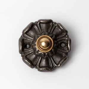 May include: A black cast iron doorbell button with a gold-toned push button in the center. The button has a decorative, floral design.