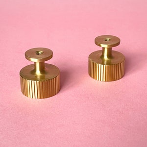 May include: Two gold-colored cabinet knobs with a ridged cylindrical base and a small, round top. The knobs are made of metal and sit on a pink background. The knobs are likely for furniture or other decorative purposes.