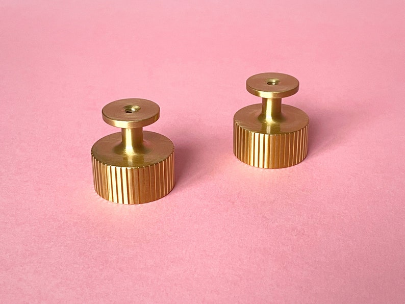 May include: Two gold-colored cabinet knobs with a ridged cylindrical base and a small, round top. The knobs are made of metal and sit on a pink background. The knobs are likely for furniture or other decorative purposes.