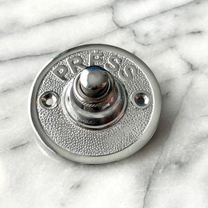 May include: A round, chrome-plated doorbell button with the word "PRESS" embossed on the face. The button has a raised, circular knob in the centre.