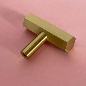 Op de afbeelding: Een goudkleurige T-vormige kastknop met een zeshoekige basis.