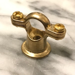 May include: A brass pipe clamp with a circular loop and two screws. The clamp has a threaded base and is designed to secure pipes or other cylindrical objects. The metal has a shiny, golden finish.