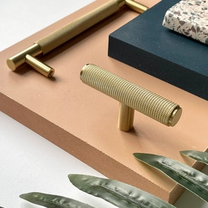 May include: Two gold metal cabinet knobs with a textured finish. The larger knob is a pull handle, and the smaller knob is a t-shaped knob.