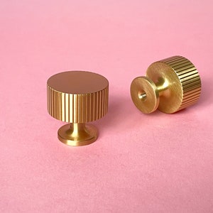May include: Two gold-colored cabinet knobs with a ridged cylindrical design and a circular top. One knob is upright, and the other is lying on its side. The knobs are on a pink background.