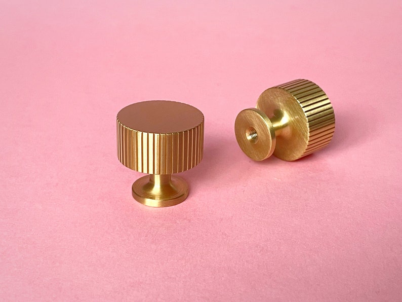 May include: Two gold-colored cabinet knobs with a ridged cylindrical design and a circular top. One knob is upright, and the other is lying on its side. The knobs are on a pink background.