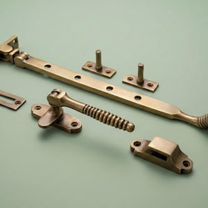 Könnte beinhalten: Eine Sammlung antiker Fensterbeschläge aus Messing, darunter ein Fensterfeststeller, ein Griff und verschiedene Montageplatten. Die Teile sind auf einer hellgrünen Oberfläche angeordnet und zeigen ihre gealterte Oberfläche und aufwendigen Designs.