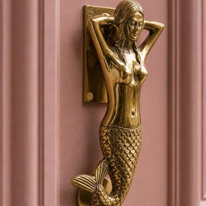 Puede incluir: Un llamador de puerta de latón pulido con forma de sirena con los brazos levantados detrás de la cabeza. La figura presenta un torso humano detallado y una cola de pez texturizada con escamas. Se fija a la puerta mediante dos placas circulares.