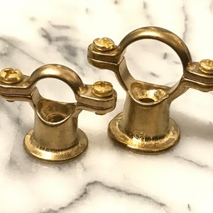 May include: Two brass pipe clamps, each with a circular top and a base. They have two screws for securing pipes. The clamps are made of shiny, golden metal and are set against a white and grey marble background.