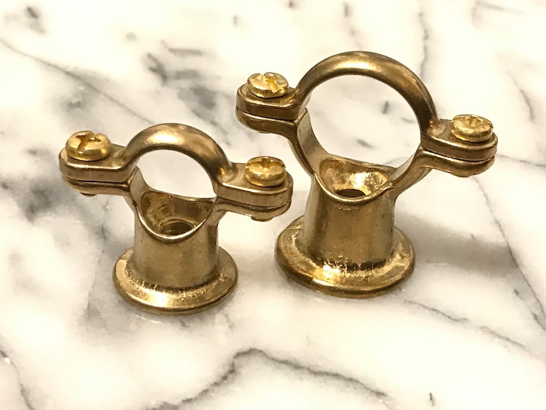 May include: Two brass pipe clamps, each with a circular top and a base. They have two screws for securing pipes. The clamps are made of shiny, golden metal and are set against a white and grey marble background.