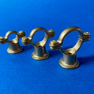 May include: Three antique brass pipe clamps with a circular design and two screws each. The clamps have a circular ring and a base, set against a blue background. These clamps are likely used for plumbing or industrial applications.
