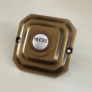 May include: Antique brass doorbell button with a white center. The button is inscribed with the word "PRESS" in black lettering. The doorbell has a square shape with rounded edges and two screw holes.