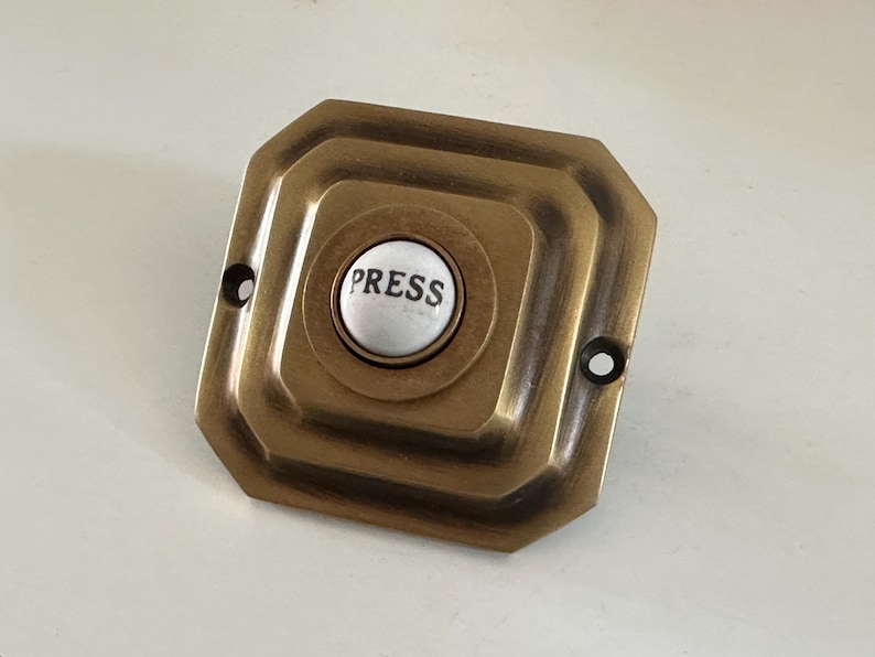 May include: Antique brass doorbell button with a white center. The button is inscribed with the word "PRESS" in black lettering. The doorbell has a square shape with rounded edges and two screw holes.