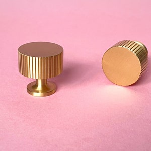 May include: Two gold-colored cabinet knobs with a ridged design. One knob is upright, showing the base and the ridged top. The other knob is on its side, displaying the ridged edge. The background is a solid pink.