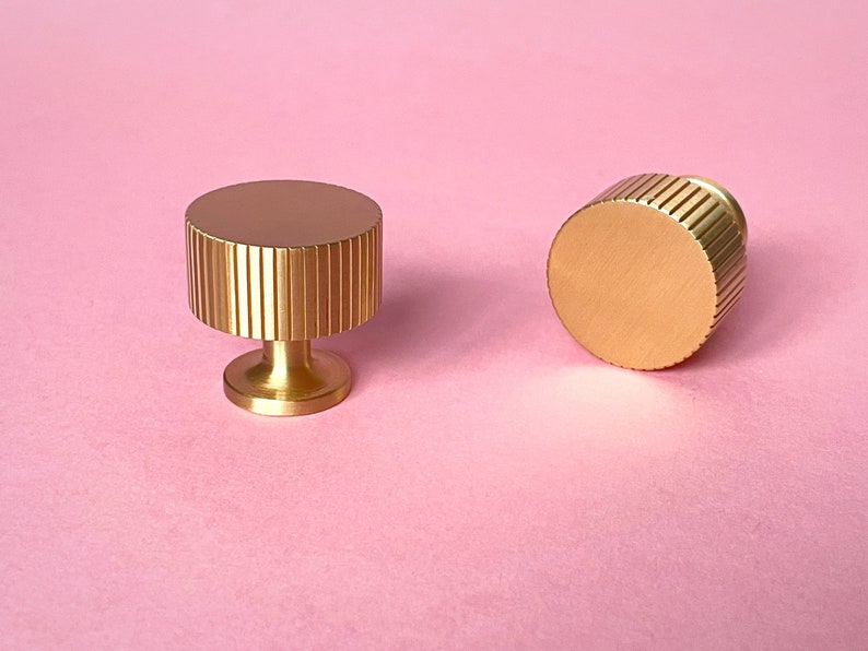 May include: Two gold-colored cabinet knobs with a ridged design. One knob is upright, showing the base and the ridged top. The other knob is on its side, displaying the ridged edge. The background is a solid pink.