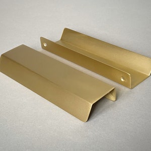 May include: Two gold metal drawer pulls with a rectangular shape and a slight curve at the top. The pulls have a brushed finish.