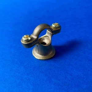 May include: A bronze-coloured metal bracket with a curved top and a circular base. Two screws secure the curved portion to the base. The bracket is set against a blue background.