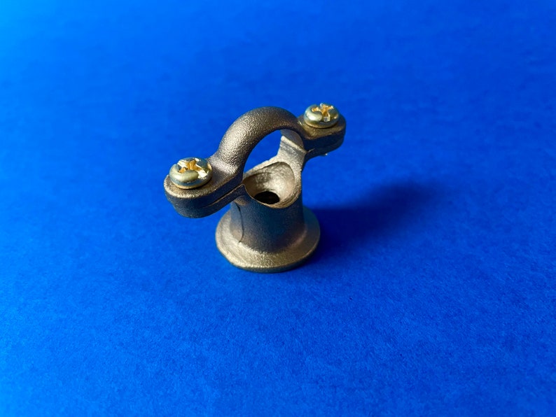 May include: A bronze-coloured metal bracket with a curved top and a circular base. Two screws secure the curved portion to the base. The bracket is set against a blue background.