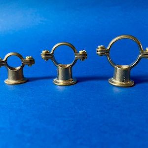 May include: Three brass pipe clamps of different sizes are displayed against a blue backdrop. Each clamp has a circular ring secured by two screws, attached to a short, cylindrical base. The clamps are made of a brass-coloured metal.