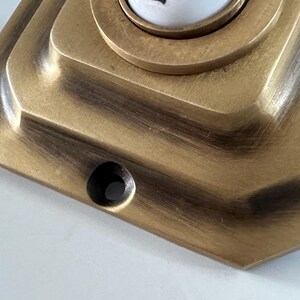 May include: Close-up of a vintage brass doorbell button with a geometric design. The button has a circular center and a rectangular base with a dark patina, suggesting age and use. The finish is a warm, antique gold.