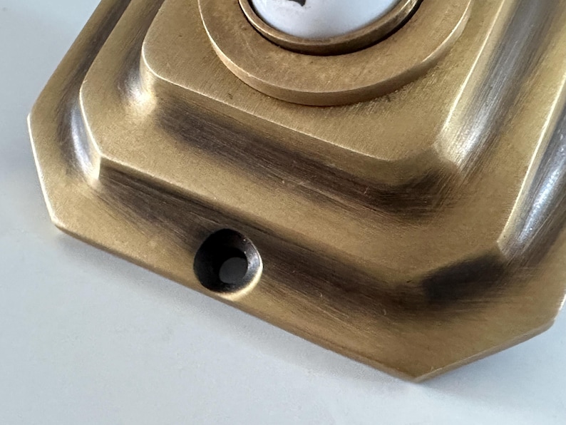 May include: Close-up of a vintage brass doorbell button with a geometric design. The button has a circular center and a rectangular base with a dark patina, suggesting age and use. The finish is a warm, antique gold.