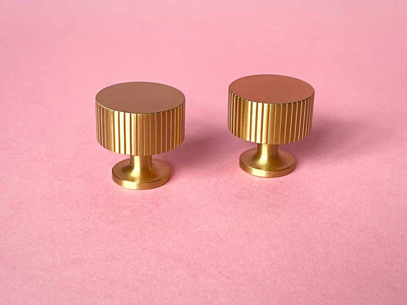May include: Two gold-colored cabinet knobs with a cylindrical, ridged design on a pink background. The knobs have a flat, circular top and a short, rounded base. The knobs are likely used for furniture or decorative purposes.