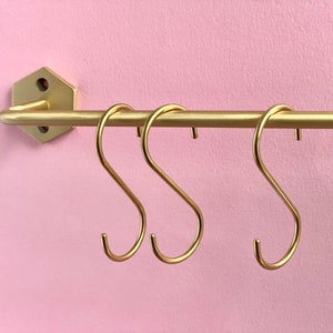 Peut inclure: Une barre métallique dorée avec trois crochets en forme de S, fixée sur un mur rose. La barre est fixée au mur avec un support hexagonal. Les crochets sont espacés uniformément.
