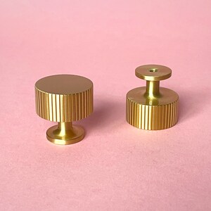 May include: Two gold-colored cabinet knobs on a pink background. The knobs have a cylindrical shape with vertical ridged detailing. One knob has a flat top, while the other has a raised, circular top with a hole. These decorative knobs are likely for furniture.