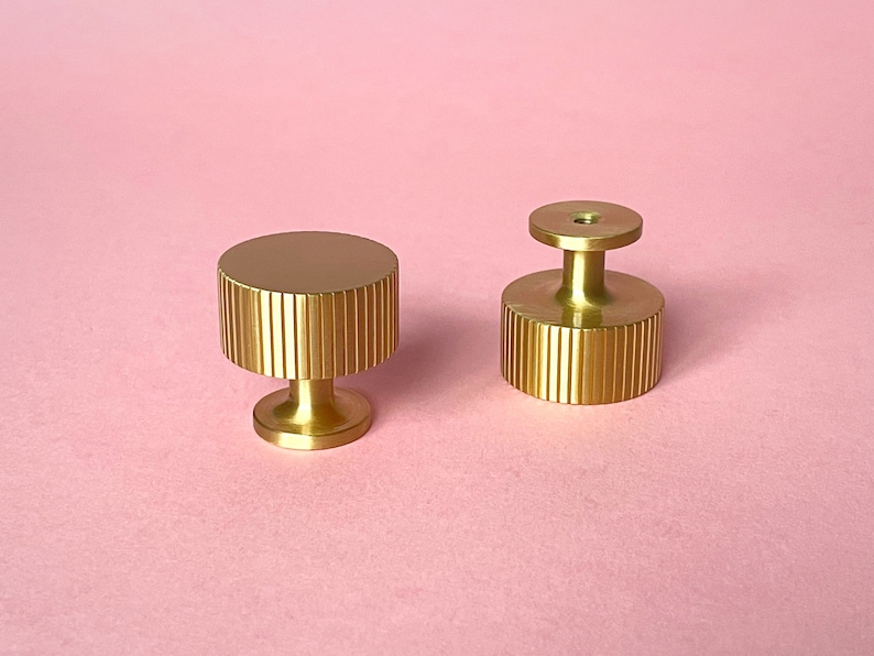 May include: Two gold-colored cabinet knobs on a pink background. The knobs have a cylindrical shape with vertical ridged detailing. One knob has a flat top, while the other has a raised, circular top with a hole. These decorative knobs are likely for furniture.