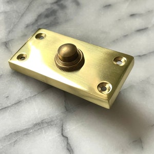 May include: A rectangular brass doorbell button with a rounded button in the centre. The button has a textured ring around the base. Four screw holes are in each corner of the rectangular plate.