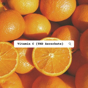 Pu&ograve; includere: Una collezione di agrumi color arancione brillante, tra cui arance intere e due arance tagliate a met&agrave; che mostrano i segmenti interni, sovrapposta a un'etichetta rettangolare bianca contenente il testo nero Vitamin C (THD Ascorbate) e un'icona a forma di lente d'ingrandimento.