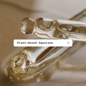 May include: Two clear glass bottles with a dropper tip filled with a light yellow liquid. The bottles are lying on a light brown surface. The text "Plant-Based Squalane" is displayed in a search bar with a magnifying glass icon.