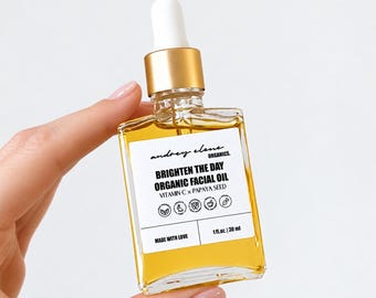 Puede incluir: Un frasco de vidrio cuadrado de aceite facial orgánico Brighten The Day con tapa de gotero dorada. La etiqueta blanca presenta texto en negro, Vitamin C x Papaya Seed y seis iconos circulares. El frasco contiene 30 ml de aceite líquido amarillo.