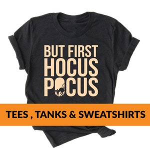 Pode incluir: Camiseta preta com o texto "But First Hocus Pocus" em laranja claro. Uma silhueta de gato preto está dentro do "O" de "Pocus". O texto está centralizado na camiseta. A camiseta está dobrada e exibida em um fundo branco. Tees, Tanks & Sweatshirts.