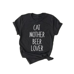 Może przedstawiać: Czarny T-shirt z białym napisem "Cat Mother Beer Lover".