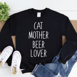 Może przedstawiać: Czarny sweter z białym napisem "Cat Mother Beer Lover". Sweter leży płasko na białym drewnianym tle z parą białych but&oacute;w sportowych i niebieskimi dżinsami.