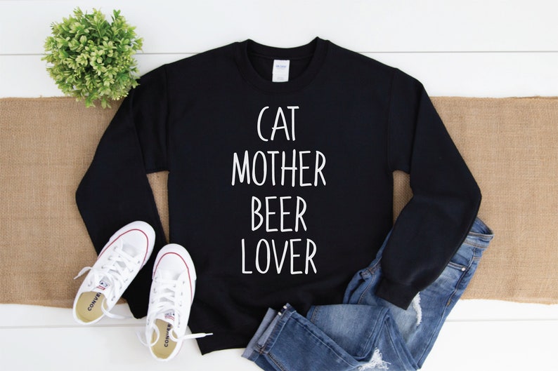 Może przedstawiać: Czarny sweter z białym napisem "Cat Mother Beer Lover". Sweter leży płasko na białym drewnianym tle z parą białych but&oacute;w sportowych i niebieskimi dżinsami.