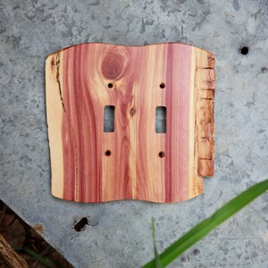 May include: A wooden double light switch cover with a natural edge. The cover is made of reddish-brown wood with visible grain and two rectangular openings for the switches. The wood has a rough, unfinished edge.