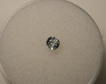 May include: A small, round, clear gemstone with a slight blue tint. The gemstone is sitting on a white, textured surface.
