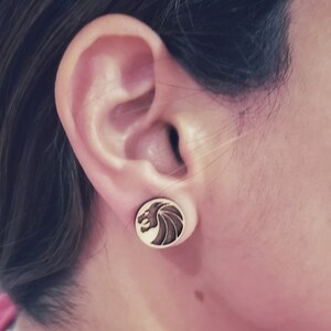 May include: Wooden stud earrings with a dragon design. The earrings are round with a light wood color and a dark brown dragon illustration. The dragon has a wing-like design and is facing left.
