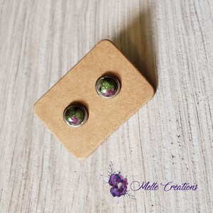 Peut inclure: Une paire de boucles d'oreilles en argent avec des accents de pierres précieuses vertes et roses. Les boucles d'oreilles sont présentées sur une carte marron.