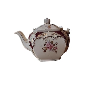 Può includere: Una teiera in ceramica color crema con un motivo a reticolo bordeaux e oro lungo il bordo superiore e il coperchio. Il corpo è decorato con un motivo floreale centrale di rose rosa, bordi dorati e un beccuccio curvo con coperchio.