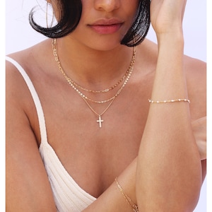 May include: A layered gold necklace with a cross pendant, paired with a delicate bracelet. The jewelry is worn on a person wearing a white tank top. The necklace features a variety of chain styles, creating a stylish and elegant look.