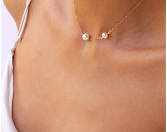 May include: A delicate gold necklace featuring two white pearls. The necklace is displayed on a person's neck, with a white top visible in the background. The pearls are round and evenly spaced on the gold chain.