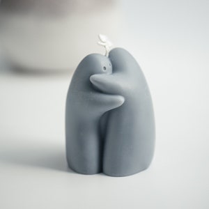 Peut inclure: Une bougie grise en forme de deux personnages enlacés. La bougie a une texture lisse et une petite mèche. En arrière-plan, un vase flou.