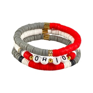 Peut inclure: Un ensemble de trois bracelets avec des perles rouges, blanches et grises. Les bracelets sont empilés les uns sur les autres et épellent le mot "OHIO" en lettres noires sur des perles blanches.
