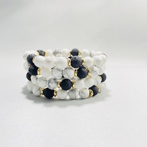 Puede incluir: Un conjunto de cuatro pulseras hechas con cuentas de howlite blanca y lava negra. Cada pulsera tiene separadores dorados entre las cuentas.