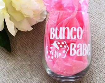 Puede incluir: Una copa de vino de vidrio transparente con una calcomanía de vinilo blanco que dice "Bunco Babe" con un par de dados. La copa está sobre papel de seda rosa.